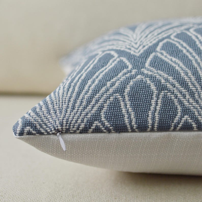May include: Close-up of a decorative pillow with a blue and white geometric pattern. The pillow has a white zipper and a neutral-colored base. The pattern is a repeating design of white lines on a blue background. The pillow is made of a textured fabric.
