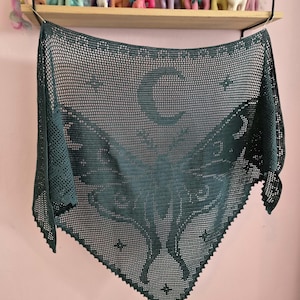 May include: A dark green triangular crochet shawl featuring an intricate moth design at the centre, a crescent moon motif above the moth, and small star patterns surrounding it. The shawl has a mesh-like texture with a decorative picot edge.