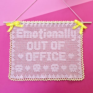 Puede incluir: Un tapiz rectangular de ganchillo rosa claro con el texto "SMILE OUT OF FOCUS" en un tono rosa más oscuro. La pieza cuelga de un palo de madera con una cuerda rosa y está decorada con dos lazos amarillos de lunares en las esquinas superiores.