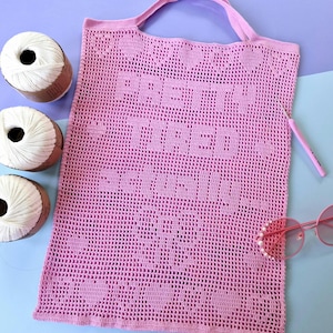 May include: A light pink crochet tote bag featuring the text "PRETTY TIRED SERIOUSLY" integrated into the mesh pattern. The bag includes two top handles and is accompanied by three white yarn balls, a pink crochet hook, and pink sunglasses.
