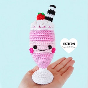 Puede incluir: Un batido de fresa de ganchillo rosa y blanco con una cara sonriente, una fresa roja y una pajita a rayas blancas y negras. El batido está sentado sobre una base de ganchillo blanca. El texto "PATTERN Super Cute Design" está en un corazón blanco.