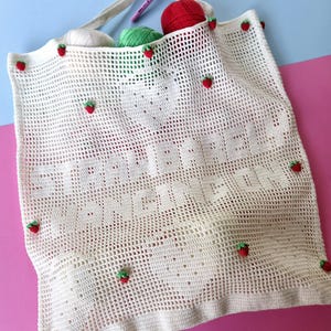 Peut inclure: Un sac fourre-tout en crochet de couleur crème avec un motif en grille, de petites fraises rouges appliquées et le texte "STRAWBERRY HANGING ON" intégré au design. Le sac contient trois pelotes de laine blanche, verte et rouge et un crochet rose.
