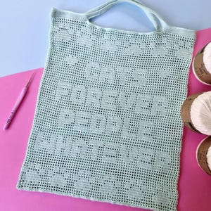 May include: A light mint green crocheted tote bag featuring a grid pattern and the text "CATS FOREVER PEOPLE NEVER" integrated into the design. The bag includes two handles and is displayed alongside a pink crochet hook and three white yarn balls.