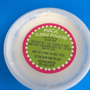May include: A white plastic container with a pink and green label that reads "MAGIC BUBBLE POWDER" and includes instructions for making giant bubbles. The label also includes the text "MADE WITH GUAR GUM AND BAKING POWDER" and a phone number and email address.