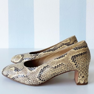 May include: A pair of vintage snakeskin-patterned pumps. The shoes have a neutral color palette with black and tan tones. The heels are a block style, and the shoes have a rounded toe. The background is a white surface with blue and white stripes.