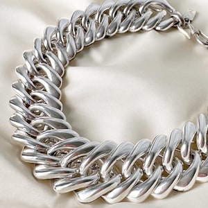 May include: A chunky silver-toned chain necklace featuring large, polished, interlocking oval links. The necklace has a high-shine metallic finish and includes a small rectangular metal tag attached near the clasp mechanism on the right side.