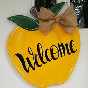 Op de afbeelding: Een geel, appelvormig houten deurbord met het woord "Welcome" in zwart schrift. Het heeft groene bladeren, een bruine steel en een strik van jute. Het bord hangt aan een witte deur.