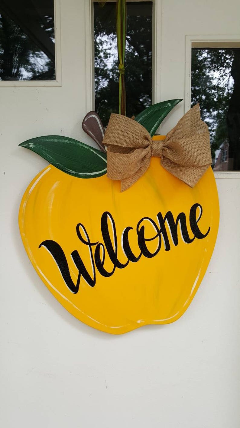 Op de afbeelding: Een geel, appelvormig houten deurbord met het woord "Welcome" in zwart schrift. Het heeft groene bladeren, een bruine steel en een strik van jute. Het bord hangt aan een witte deur.