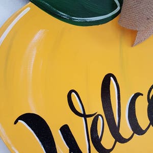 Op de afbeelding: Een houten welkomstbord in de vorm van een citroen. Het bord is geel geschilderd met een groen blad en een bruine steel. Het woord "Welkom" staat in zwarte letters op de voorkant. Een jute strik is aan de bovenkant bevestigd.
