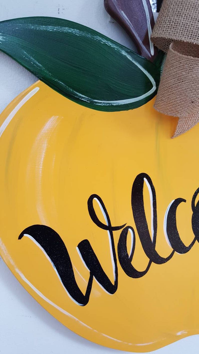 Op de afbeelding: Een houten welkomstbord in de vorm van een citroen. Het bord is geel geschilderd met een groen blad en een bruine steel. Het woord "Welkom" staat in zwarte letters op de voorkant. Een jute strik is aan de bovenkant bevestigd.