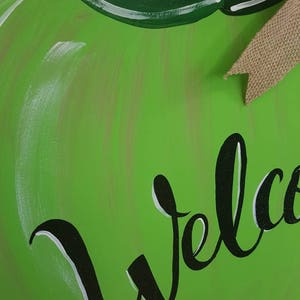 Op de afbeelding: Een groen, appelvormig houten bord met het woord "Welcome" in zwart schrift. Het bord heeft een jute strik en groene bladeren. De appel is lichtgroen geschilderd.