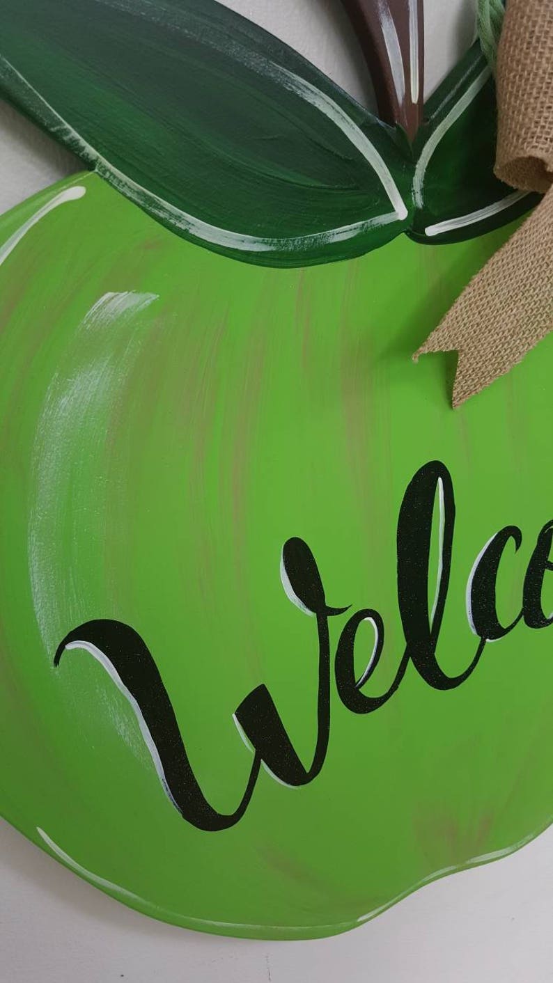 Op de afbeelding: Een groen, appelvormig houten bord met het woord "Welcome" in zwart schrift. Het bord heeft een jute strik en groene bladeren. De appel is lichtgroen geschilderd.