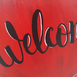 Op de afbeelding: Een rood, houten hartvormig bord met het woord "Welcome" in zwart, glitterig script. Het bord heeft een gladde, glanzende afwerking en is geschikt voor woondecoratie.