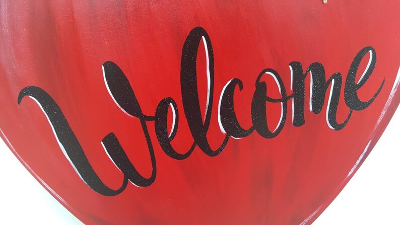 Op de afbeelding: Een rood, houten hartvormig bord met het woord "Welcome" in zwart, glitterig script. Het bord heeft een gladde, glanzende afwerking en is geschikt voor woondecoratie.