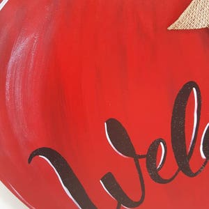 Op de afbeelding: Een houten appelvormig bord met een helderrode basis, groene bladeren en een jute strik. Het woord "Welcome" is geschreven in een cursief lettertype met een glitteromtrek. Ontworpen voor woondecoratie.