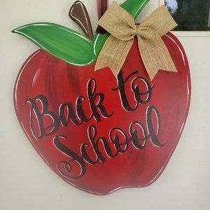 Op de afbeelding: Een houten appelvormig bord met de tekst "Back to School" in zwart script. De appel is rood met groene bladeren, een bruine steel en een strik van jute. Ontworpen om op te hangen.