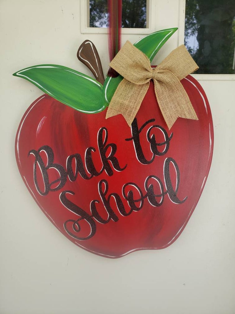 Op de afbeelding: Een houten appelvormig bord met de tekst "Back to School" in zwart script. De appel is rood met groene bladeren, een bruine steel en een strik van jute. Ontworpen om op te hangen.
