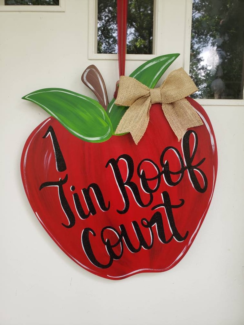 Op de afbeelding: Rode, appelvormige houten deurhanger met de tekst "1 Tin Roof Court" in zwart script. De appel heeft groene bladeren, een bruine steel en een strik van jute. De deurhanger is ontworpen voor woondecoratie.