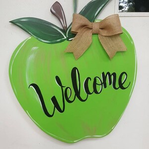 Op de afbeelding: Een groene, appelvormige houten deurhanger met het woord "Welcome" in zwart schrift. Het heeft groene bladeren, een bruine steel en een strik van jute. De deurhanger is ontworpen om aan een deur te worden gehangen.