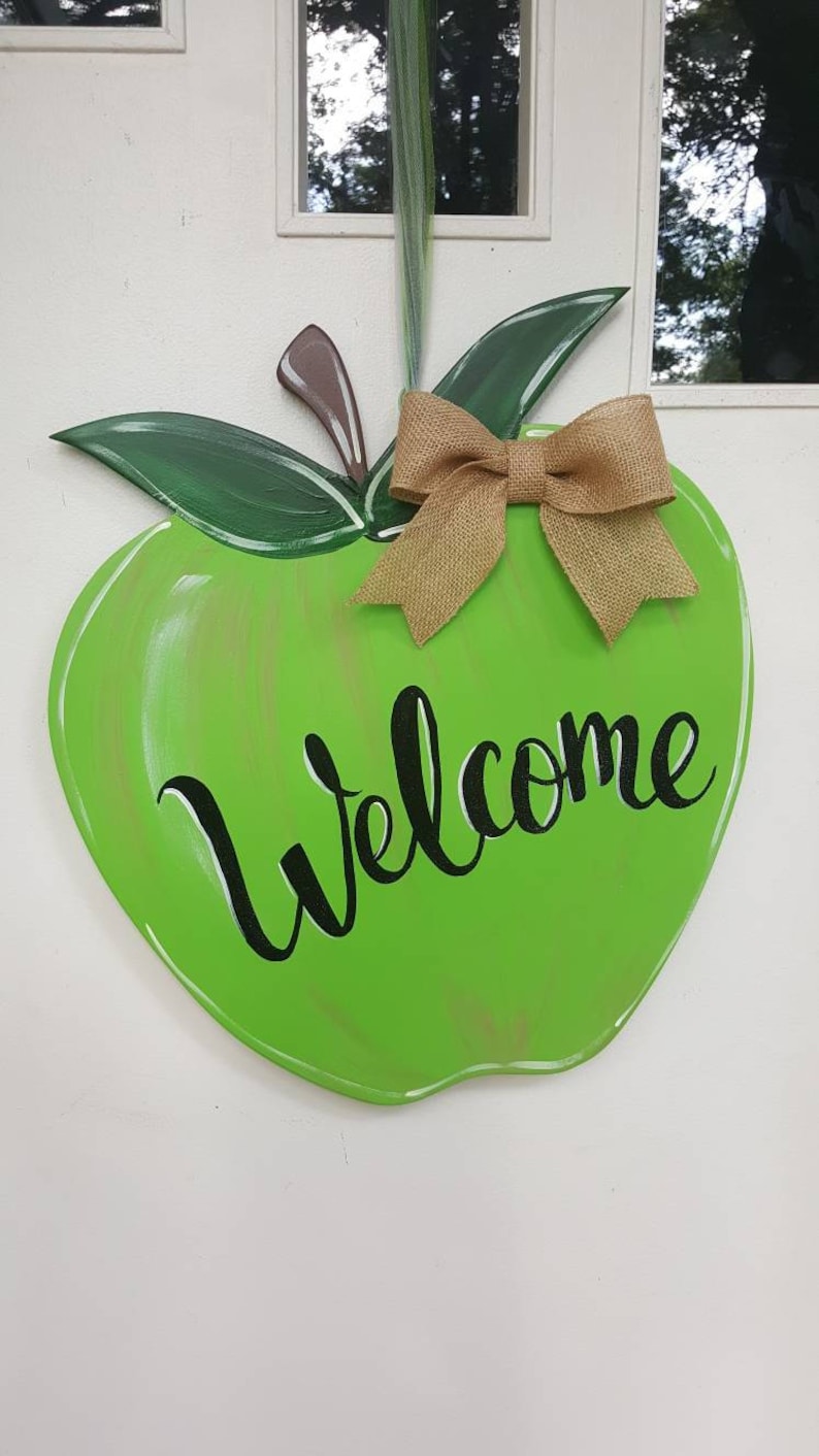 Op de afbeelding: Een groene, appelvormige houten deurhanger met het woord "Welcome" in zwart schrift. Het heeft groene bladeren, een bruine steel en een strik van jute. De deurhanger is ontworpen om aan een deur te worden gehangen.