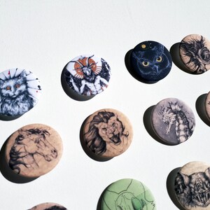K&ouml;nnte beinhalten: Eine Sammlung von 12 runden Pins mit verschiedenen Schwarzwei&szlig;-Illustrationen von Tieren, darunter eine Katze, ein L&ouml;we, ein Seepferdchen, eine Fledermaus und ein geh&ouml;rntes Wesen. Die Pins befinden sich auf einem wei&szlig;en Hintergrund.