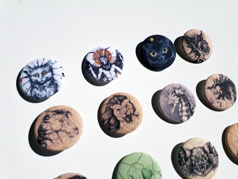 K&ouml;nnte beinhalten: Eine Sammlung von 12 runden Pins mit verschiedenen Schwarzwei&szlig;-Illustrationen von Tieren, darunter eine Katze, ein L&ouml;we, ein Seepferdchen, eine Fledermaus und ein geh&ouml;rntes Wesen. Die Pins befinden sich auf einem wei&szlig;en Hintergrund.