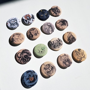 K&ouml;nnte beinhalten: Eine Sammlung von 16 runden Pins mit verschiedenen Schwarzwei&szlig;-Illustrationen von Tieren, darunter eine Katze, ein Seepferdchen, ein Wolf, ein L&ouml;we und ein Sch&auml;del. Die Pins sind in einem Raster auf einer wei&szlig;en Oberfl&auml;che angeordnet.