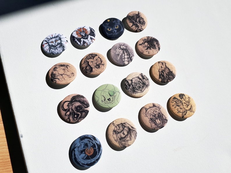 K&ouml;nnte beinhalten: Eine Sammlung von 16 runden Pins mit verschiedenen Schwarzwei&szlig;-Illustrationen von Tieren, darunter eine Katze, ein Seepferdchen, ein Wolf, ein L&ouml;we und ein Sch&auml;del. Die Pins sind in einem Raster auf einer wei&szlig;en Oberfl&auml;che angeordnet.