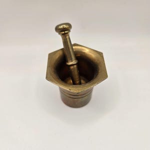 May include: A vintage brass mortar and pestle. The mortar has a hexagonal top and a cylindrical base. The pestle is cylindrical with a rounded head. The set is made of brass and has a warm, antique finish.