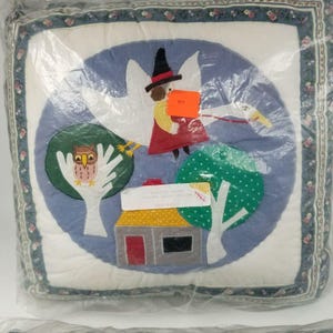 Puede incluir: Un cojín cuadrado con un diseño caprichoso que presenta una bruja montada en una cigüeña, un búho en un árbol y una casa. El cojín tiene una combinación de colores azul y blanco con un borde floral y está envuelto en plástico.