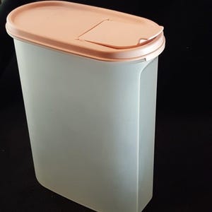 Puede incluir: Un recipiente alto de plástico translúcido de forma ovalada con una tapa de color melocotón. La tapa tiene una pequeña abertura con bisagras. El recipiente está diseñado para el almacenamiento de alimentos.