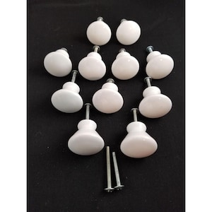 May include: A collection of eleven white ceramic cabinet knobs with silver hardware. The knobs are arranged on a black surface, with two additional screws visible. The knobs have a smooth, rounded design, suitable for furniture or decorative use.