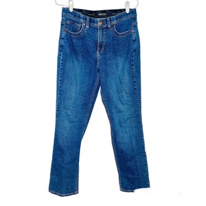 Puede incluir: Vaqueros de mezclilla azules con cintura alta y un diseño clásico de cinco bolsillos. Los vaqueros están colgados de una percha blanca. El denim tiene un lavado medio con detalles sutiles de desvanecimiento y bigotes.