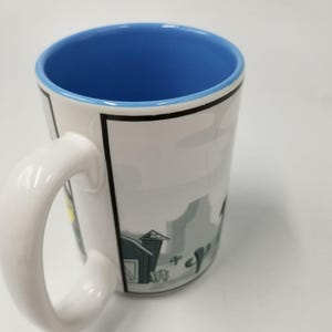 May include: White ceramic mug with a blue interior and a white handle. The mug features a black border and a cartoon illustration of a desert scene with a barn and cacti. The mug is designed for hot beverages.