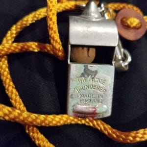 May include: Close-up of a vintage metal whistle with the text "The Acme Thunderer Made in England." The whistle has a small wooden ball inside and is attached to a yellow rope. A brown button is also visible.