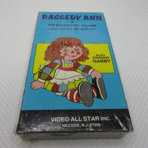 Puede incluir: Una caja de cinta VHS con Raggedy Ann, una muñeca de trapo de pelo rojo, sentada sobre un fondo azul. El título dice "Raggedy Ann en La Plaza Encantada" con el lema "¿Tienes algún castillo?" y "También protagonizada por Gabby". La caja es de Video All Star Inc. en Hillside, NJ 07205.