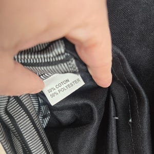 May include: A close-up of a black garment with a white label. The label reads "50% COTON 50% POLYESTER". The interior lining is a black and white striped pattern.