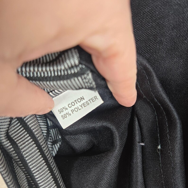 May include: A close-up of a black garment with a white label. The label reads "50% COTON 50% POLYESTER". The interior lining is a black and white striped pattern.
