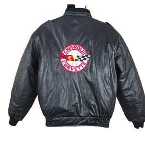Könnte beinhalten: Schwarze Jacke im Vintage-Stil mit glänzendem Finish. Auf der Rückseite befindet sich ein runder Aufnäher mit dem Chevrolet Corvette-Logo in Rot, Weiß und Schwarz. Die Jacke hat einen gerippten Kragen und Bündchen.