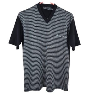 May include: A black and gray striped V-neck t-shirt with black short sleeves. The shirt has the Versace logo on the collar and a signature on the front. The shirt is hanging on a pink hanger.
