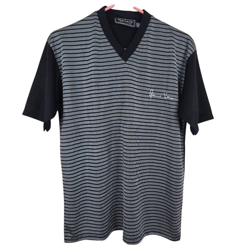 May include: A black and gray striped V-neck t-shirt with black short sleeves. The shirt has the Versace logo on the collar and a signature on the front. The shirt is hanging on a pink hanger.