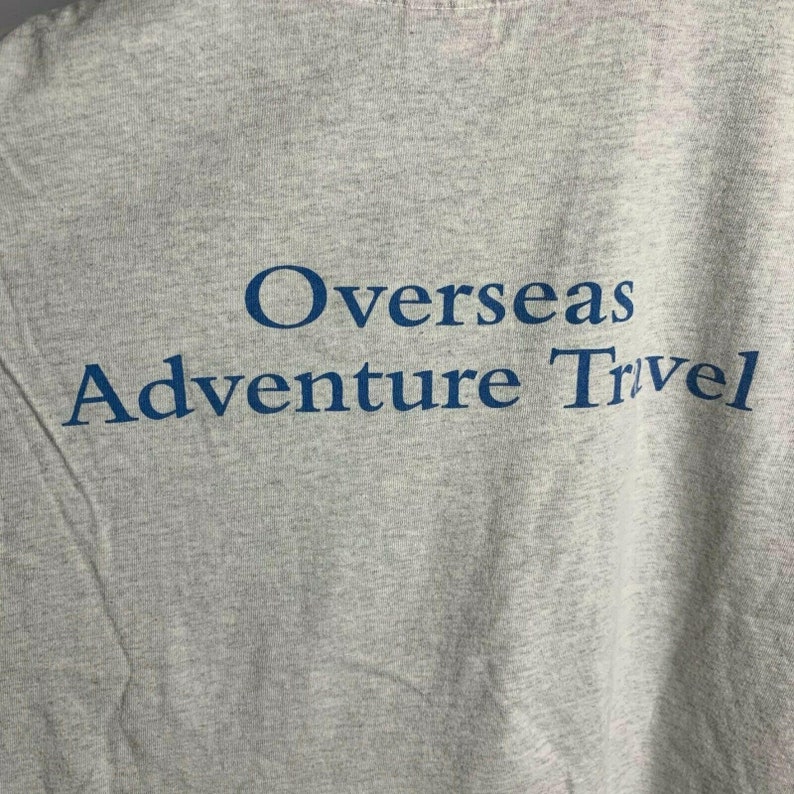 Pode incluir: Camiseta cinza com o texto "Overseas Adventure Travel" impresso em azul.