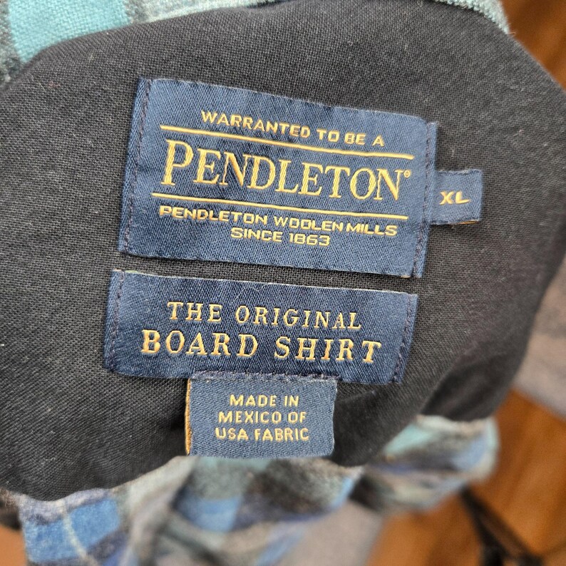 Puede incluir: Primer plano de la etiqueta de una camisa Pendleton Board. La etiqueta azul marino dice "WARRANTED TO BE A PENDLETON PENDLETON WOOLEN MILLS SINCE 1863" y "THE ORIGINAL BOARD SHIRT". La camisa es XL y est&aacute; hecha en M&eacute;xico con tela de EE. UU.
