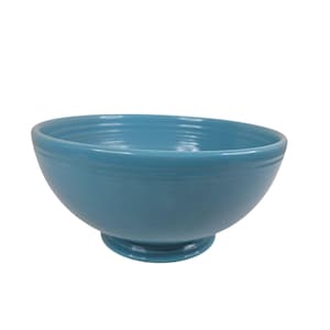 Puede incluir: Un cuenco de cerámica turquesa con base de pedestal. El cuenco tiene forma redondeada con un borde ligeramente ensanchado y un acabado liso y brillante. El cuenco es de color azul claro liso.