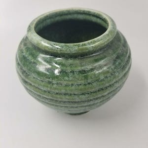 Puede incluir: Una maceta de cerámica verde con cuerpo redondeado y boca ancha. La maceta tiene una superficie texturizada y acanalada con un esmalte moteado verde claro. El borde es liso y ligeramente elevado. Esta maceta decorativa es adecuada para la decoración del hogar.