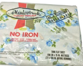 Puede incluir: Una sábana plana doble en un paquete de plástico. La sábana es blanca con un estampado floral de rosas azules y hojas verdes. El embalaje incluye el texto "Meadowbrook Luxury Percale" y "No Iron". La sábana es para un colchón de 99 x 190 cm.
