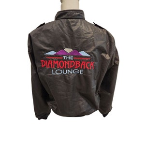 May include: A brown bomber jacket with a red and white embroidered logo that says "The Diamondback Lounge".