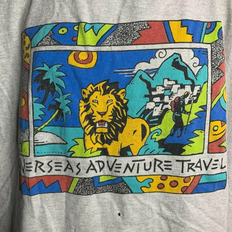 Pode incluir: Uma camiseta vintage com um design gr&aacute;fico colorido que apresenta um le&atilde;o, montanhas e palmeiras. O texto "Overseas Adventure Travel" est&aacute; impresso na parte inferior do gr&aacute;fico.