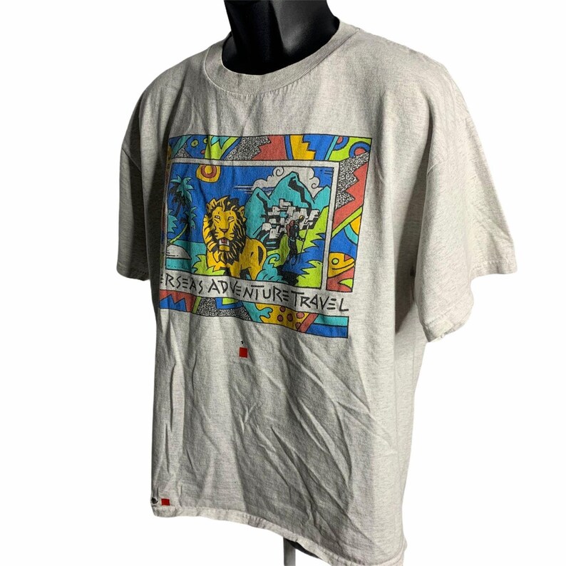 Pode incluir: Uma camiseta cinza com uma estampa gr&aacute;fica colorida de um le&atilde;o, palmeiras, montanhas e o texto "Overseas Adventure Travel".