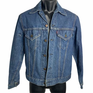 Op de afbeelding: Een klassieke blauwe denimjas met een knoopsluiting en lange mouwen. De jas heeft een puntige kraag, twee borstzakken en twee voorzakken.